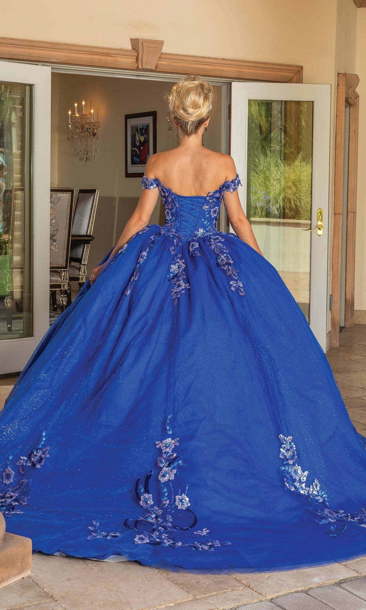 A woman stands with her back to the camera in front of open glass doors, wearing the Quinceanera Dress 1737 by Dancing Queen—a royal blue ball gown adorned with floral patterns.